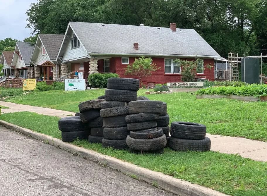 A large curbside stack of used tires in front of a residential block and community garden.