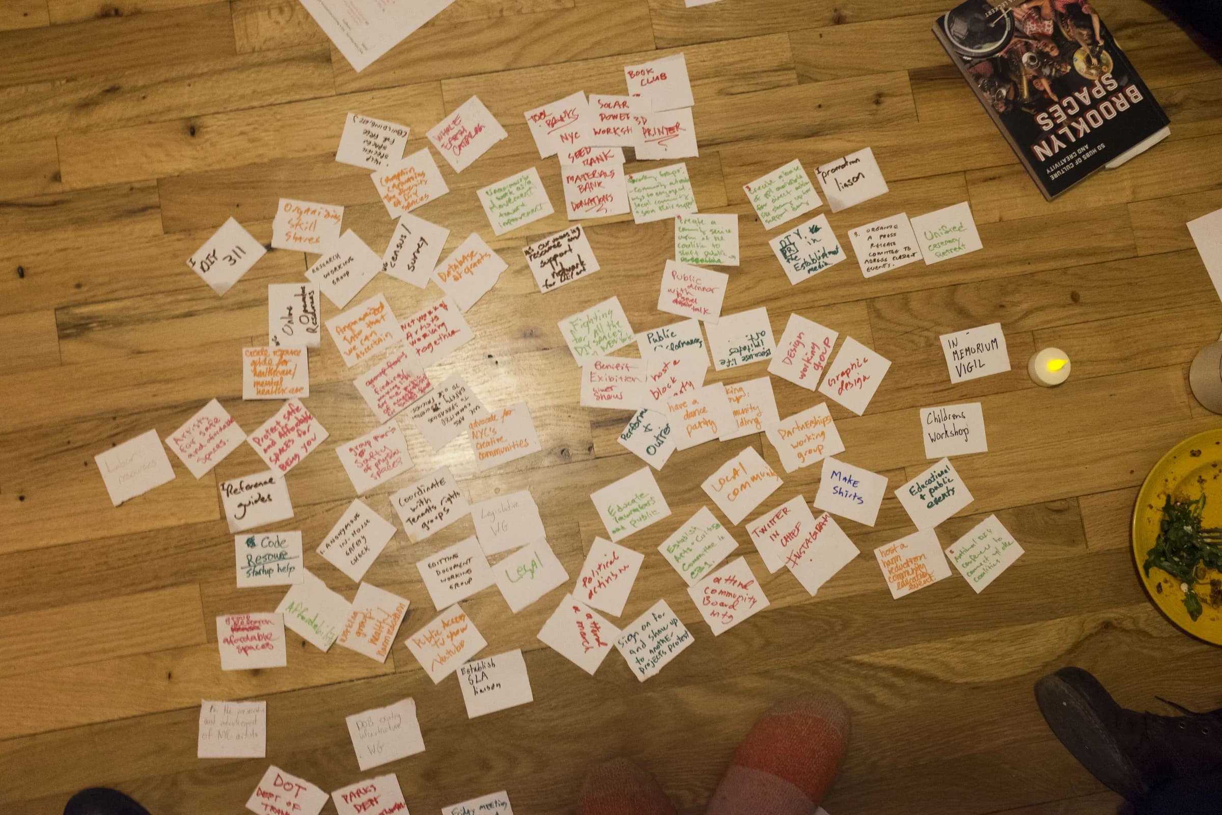 Handwritten cards in several ink colors form clusters across a wooden floor, surrounded by shoes, a book, a plate, and a small light.