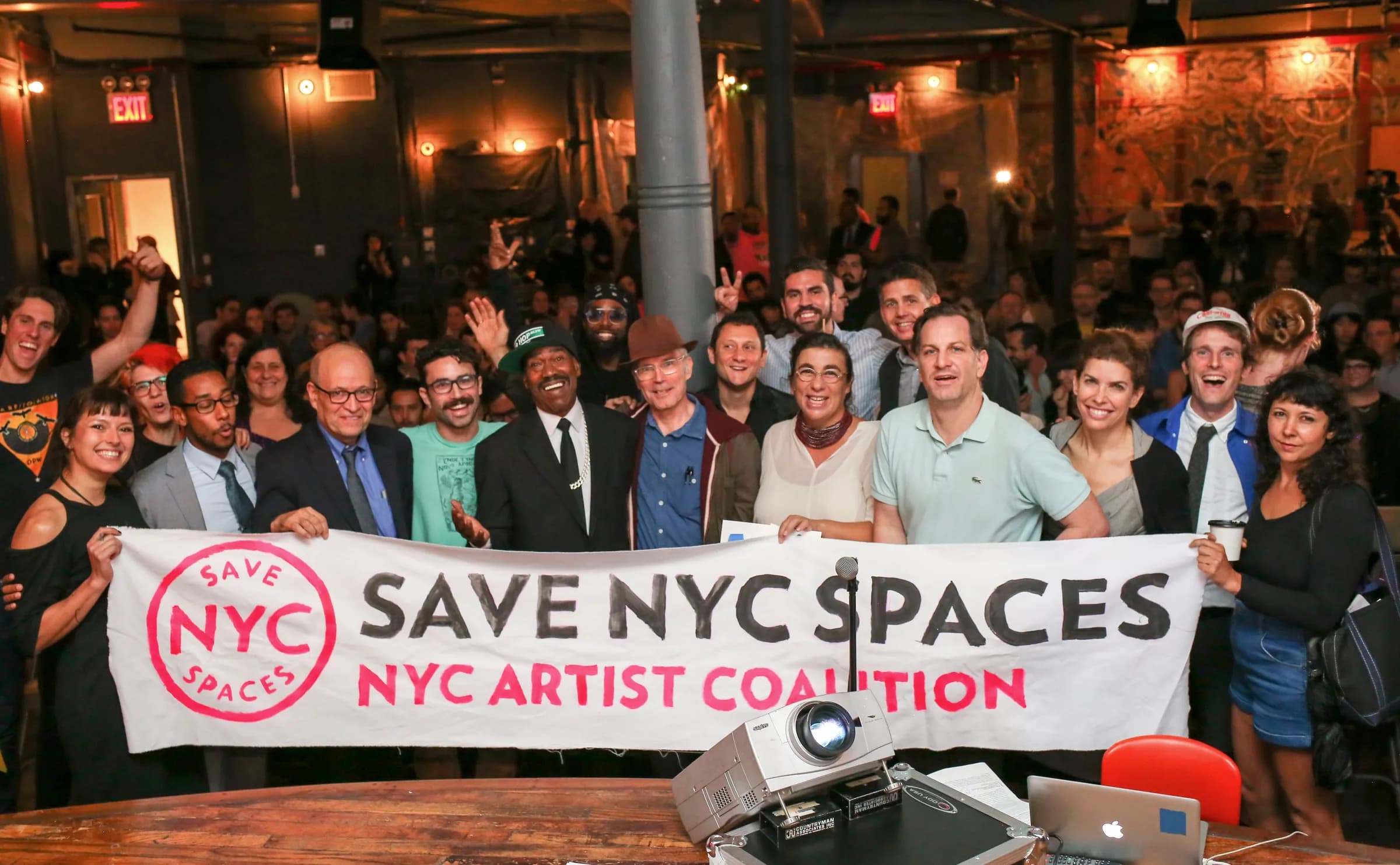 Organizers, artists, and public officials hold a hand-painted Save NYC Spaces and NYC Artist Coalition banner in front of a packed Market Hotel room.