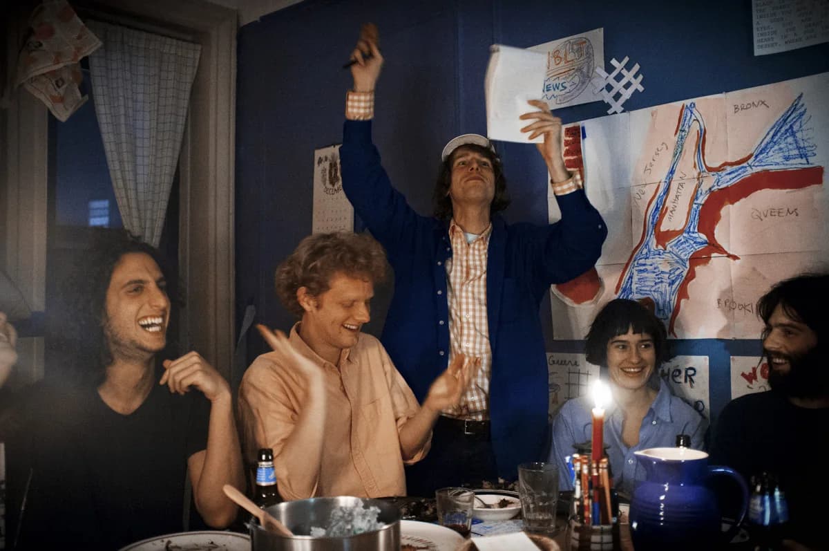 A person stands at a crowded dinner table, raising one hand and holding up a sheet of paper as others laugh beside a hand-drawn map.