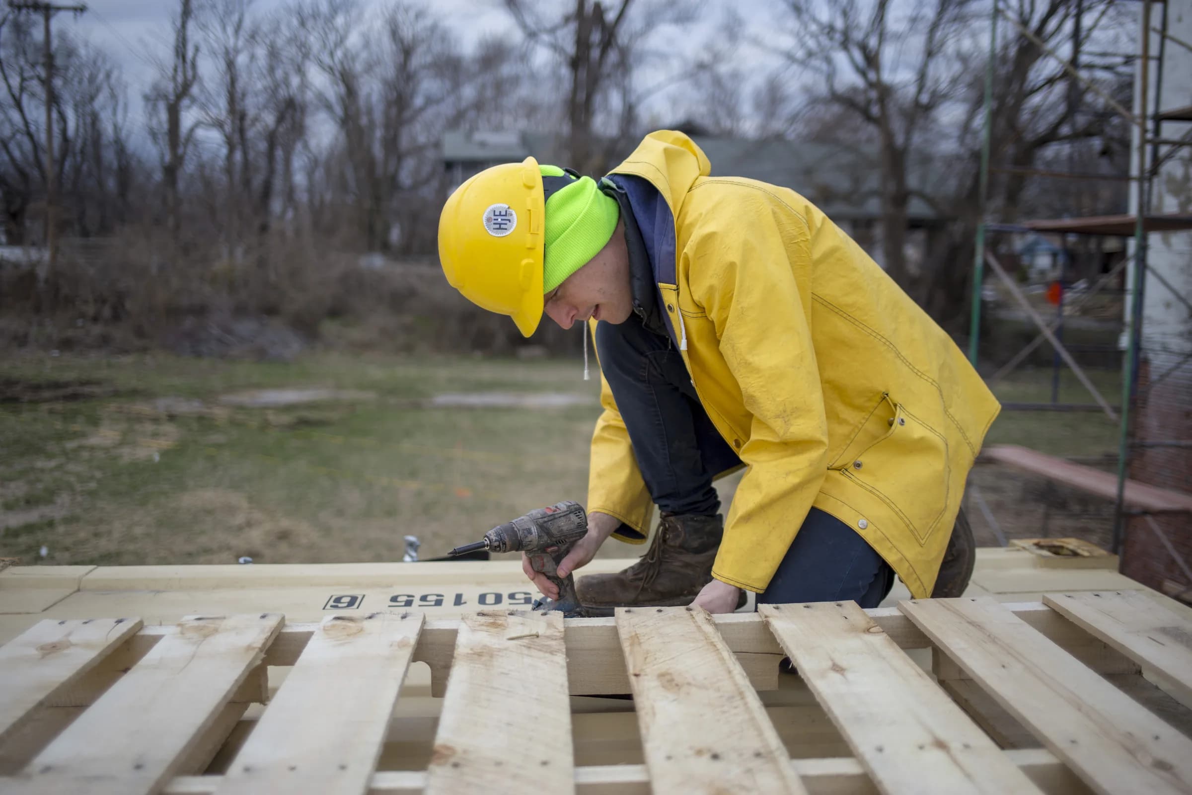 A person in a yellow hard hat and rain jacket uses a drill on a roof assembly.